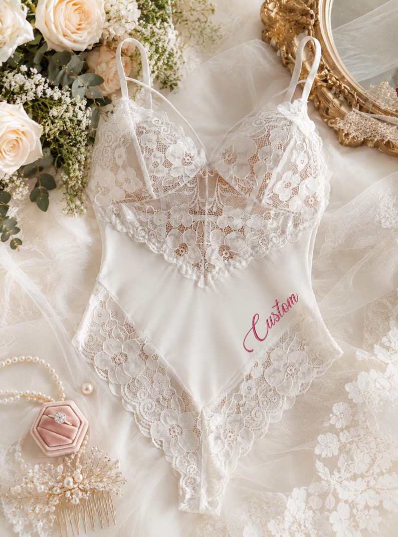 May include: A white bodysuit featuring intricate floral lace detailing on the bodice and leg openings, thin spaghetti straps, and the word Custom embroidered in a script font on the lower right side of the smooth fabric panel.