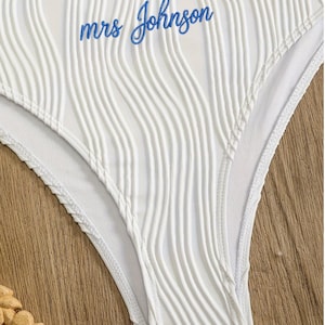 May include: White bikini bottom featuring a textured, vertical ribbed fabric pattern and a ruffled top edge. The text "mrs Johnson" is embroidered in blue cursive script across the front centre of the garment, which has a high-cut leg design.