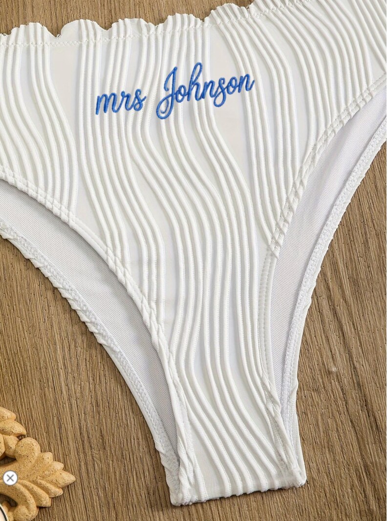 May include: White bikini bottom featuring a textured, vertical ribbed fabric pattern and a ruffled top edge. The text "mrs Johnson" is embroidered in blue cursive script across the front centre of the garment, which has a high-cut leg design.