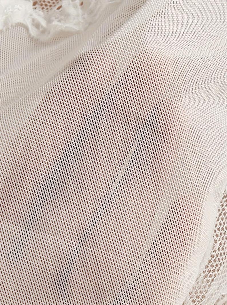 May include: A sheer white mesh fabric with a fine grid pattern. The material is translucent, revealing a subtle texture throughout the surface. Along the top and bottom edges, there is a decorative white lace trim with a scalloped floral design.