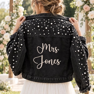 May include: A black denim jacket featuring white pearl beads and clear crystal rhinestones scattered across the back and sleeves. The back of the jacket is embroidered with the text Mrs. Jones in a light beige script font.