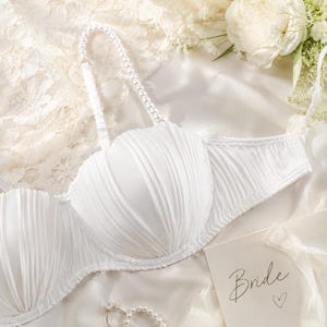 May include: A white strapless bra featuring pleated fabric cups and a single detachable shoulder strap adorned with white pearls. A small card with the word Bride and a heart symbol is visible, along with a pearl ring resting on white fabric.