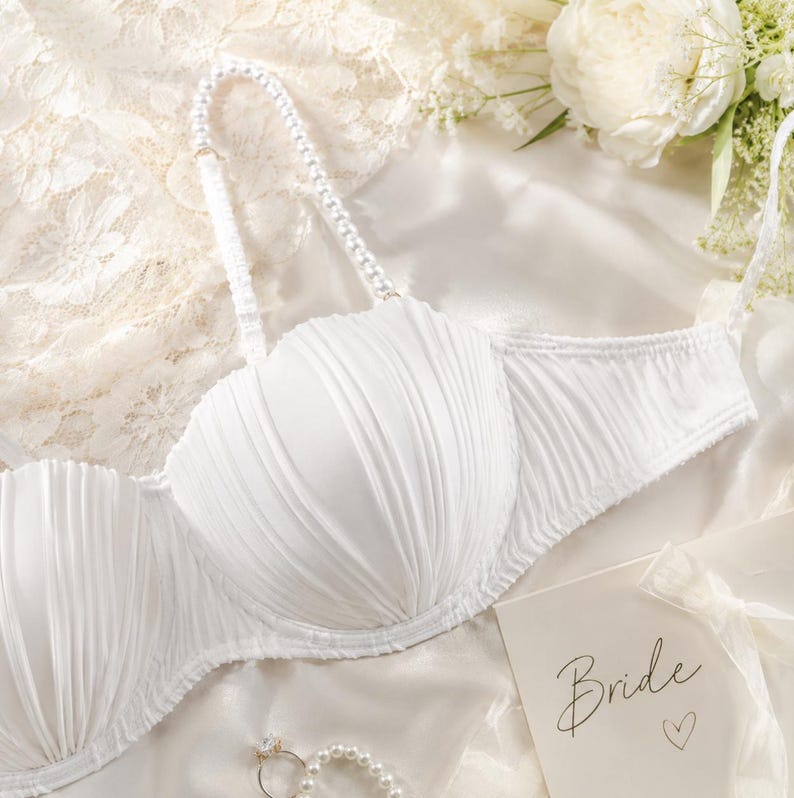 May include: A white strapless bra featuring pleated fabric cups and a single detachable shoulder strap adorned with white pearls. A small card with the word Bride and a heart symbol is visible, along with a pearl ring resting on white fabric.
