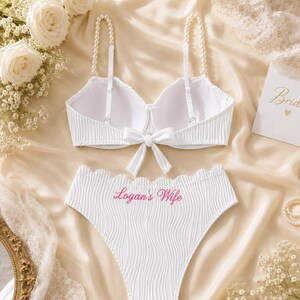 May include: A white two-piece lingerie set featuring a ribbed texture. The bra has pearl-beaded straps and a centre bow. The high-waisted briefs have a scalloped top edge and the pink embroidered text Logan's Wife centred on the front.