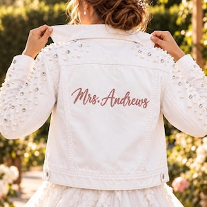 May include: A white denim jacket featuring scattered white pearls and clear rhinestones across the shoulders and sleeves. The back of the jacket displays the embroidered text Mrs. Andrews in a dusty rose script font, with a classic collar and buttons.