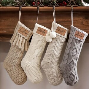 May include: Four personalized Christmas stockings in cream, off-white, and gray, hanging from a wooden mantel. Each stocking has a name tag: William, Hadley, Charlotte, and Aiden. The stockings have different knit patterns and textures.