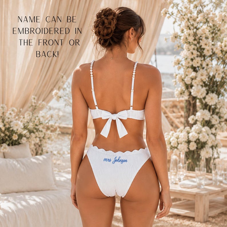 May include: A white ribbed two-piece bikini featuring thin straps with small white beads and a bow detail on the back of the top. The bikini bottom is embroidered with the blue text "mrs Johnson". Text on image: NAME CAN BE EMBROIDERED IN THE FRONT OR BACK!