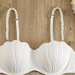 May include: A white strapless-style bra featuring a textured, vertical ribbed pattern across the cups and two shoulder straps adorned with a string of white faux pearls. The garment has a scalloped top edge and a structured, moulded cup design.