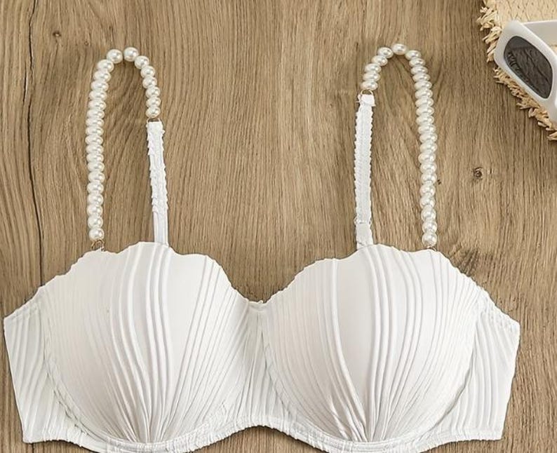 May include: A white strapless-style bra featuring a textured, vertical ribbed pattern across the cups and two shoulder straps adorned with a string of white faux pearls. The garment has a scalloped top edge and a structured, moulded cup design.