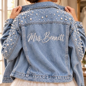May include: A light blue denim jacket featuring white pearl embellishments scattered across the back yoke and down the sleeves. The back of the jacket displays the white embroidered text "Mrs. Bennett" in a cursive script font with metal buttons.