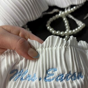 May include: A white pleated fabric garment featuring a scalloped edge and the embroidered text "Mrs. Eaton" in light blue thread. The item is accented with a string of white faux pearls, showcasing a textured, vertical ribbed design throughout.