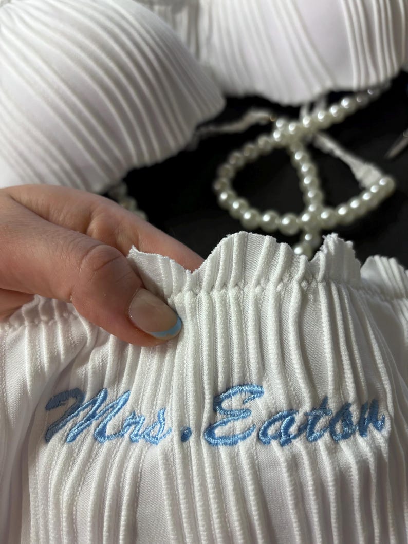 May include: A white pleated fabric garment featuring a scalloped edge and the embroidered text "Mrs. Eaton" in light blue thread. The item is accented with a string of white faux pearls, showcasing a textured, vertical ribbed design throughout.