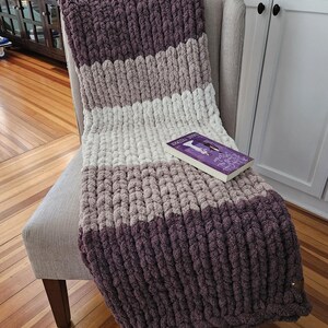 Pode incluir: Uma manta de malha grossa em tons de roxo, cinza e branco est&aacute; sobre uma cadeira. A manta tem um padr&atilde;o listrado e uma apar&ecirc;ncia macia e texturizada. Um livro com capa roxa est&aacute; sobre a manta. Um item de decora&ccedil;&atilde;o aconchegante.