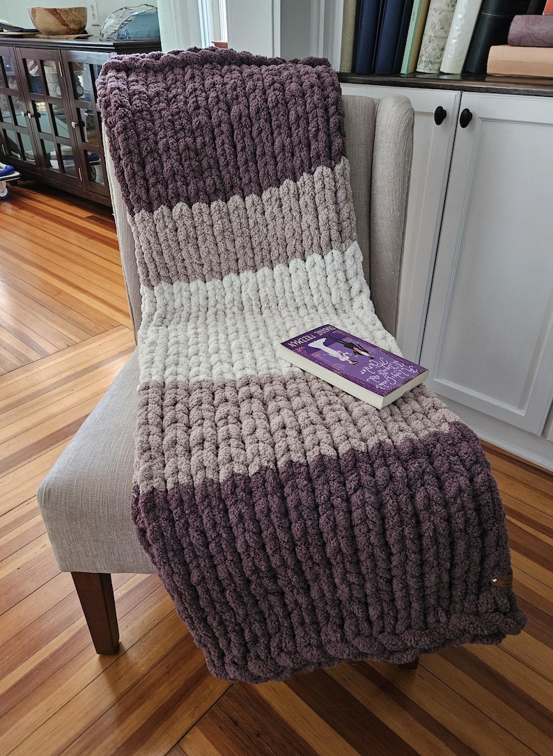 Pode incluir: Uma manta de malha grossa em tons de roxo, cinza e branco est&aacute; sobre uma cadeira. A manta tem um padr&atilde;o listrado e uma apar&ecirc;ncia macia e texturizada. Um livro com capa roxa est&aacute; sobre a manta. Um item de decora&ccedil;&atilde;o aconchegante.