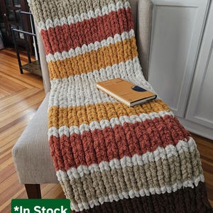 Könnte beinhalten: Eine grob gestrickte Decke in Braun-, Beige-, Orange-, Gelb- und Weißtönen liegt über einem Stuhl. Ein geschlossenes Buch mit braunem Einband liegt auf der Decke. Die Decke hat ein Streifenmuster.