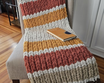 Pode incluir: Uma manta de malha grossa listrada em tons de marrom, bege, laranja, amarelo e branco está sobre uma cadeira. Um livro fechado com uma capa marrom está sobre a manta. A manta tem um padrão listrado.