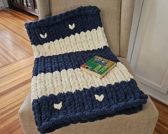 Pode incluir: Uma manta de malha grossa listrada em azul marinho e branco com detalhes em forma de coração está sobre uma poltrona bege. Um pequeno livro com o título "Goodnight Moon" está sobre a manta.