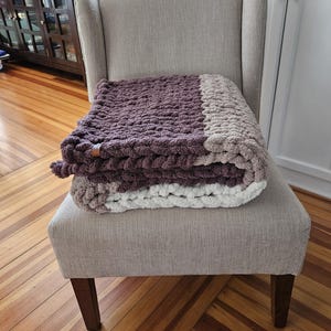 Pode incluir: Uma manta de malha grossa dobrada, em tons de roxo, cinza e branco, repousa sobre uma poltrona bege estofada. A textura da manta &eacute; vis&iacute;vel e parece macia e aconchegante. A cadeira tem encosto alto e pernas de madeira.