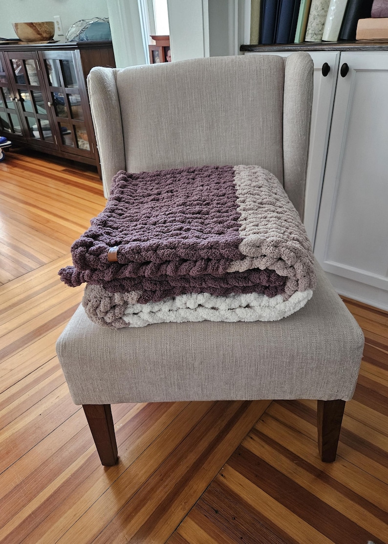 Pode incluir: Uma manta de malha grossa dobrada, em tons de roxo, cinza e branco, repousa sobre uma poltrona bege estofada. A textura da manta &eacute; vis&iacute;vel e parece macia e aconchegante. A cadeira tem encosto alto e pernas de madeira.