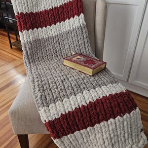 Pode incluir: Uma manta de malha grossa com listras cinza, branca e bord&ocirc; est&aacute; sobre uma poltrona bege. Um livro decorativo com p&aacute;ginas douradas descansa sobre a manta. A textura espessa da manta &eacute; vis&iacute;vel.