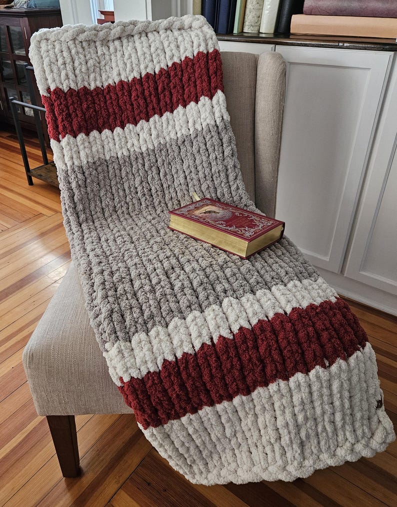 Pode incluir: Uma manta de malha grossa com listras cinza, branca e bord&ocirc; est&aacute; sobre uma poltrona bege. Um livro decorativo com p&aacute;ginas douradas descansa sobre a manta. A textura espessa da manta &eacute; vis&iacute;vel.