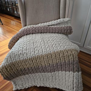 Pode incluir: Uma manta de malha grossa em tons de creme, bege e malva est&aacute; sobre uma poltrona bege. A manta apresenta listras horizontais de v&aacute;rias cores e texturas, criando uma est&eacute;tica aconchegante e convidativa. O fio grosso d&aacute; &agrave; manta uma apar&ecirc;ncia macia.