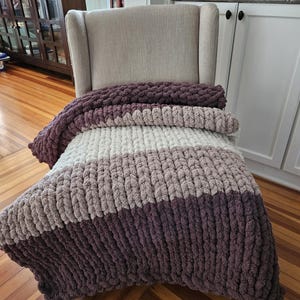 Pode incluir: Uma manta de malha grossa em tons de roxo, cinza e creme repousa sobre uma poltrona bege. A manta apresenta um design listrado com tons variados de cores. A manta aconchegante &eacute; feita de fio grosso e macio.