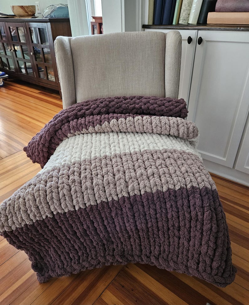 Pode incluir: Uma manta de malha grossa em tons de roxo, cinza e creme repousa sobre uma poltrona bege. A manta apresenta um design listrado com tons variados de cores. A manta aconchegante &eacute; feita de fio grosso e macio.