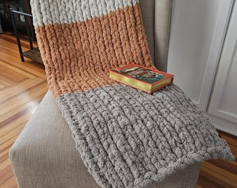 May include: A three-tone chunky knit blanket in light grey, orange, and dark grey, draped over a beige armchair. A book rests on the blanket. The blanket's texture is prominent, suggesting a soft and cosy feel, perfect for home decor.