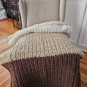 Pode incluir: Uma manta de malha grossa em tons de bege e castanho est&aacute; sobre uma poltrona bege. A manta apresenta um design texturizado e canelado, com uma sec&ccedil;&atilde;o superior bege claro, uma sec&ccedil;&atilde;o m&eacute;dia bege m&eacute;dio e uma sec&ccedil;&atilde;o inferior castanho escuro. A manta parece macia e aconchegante.