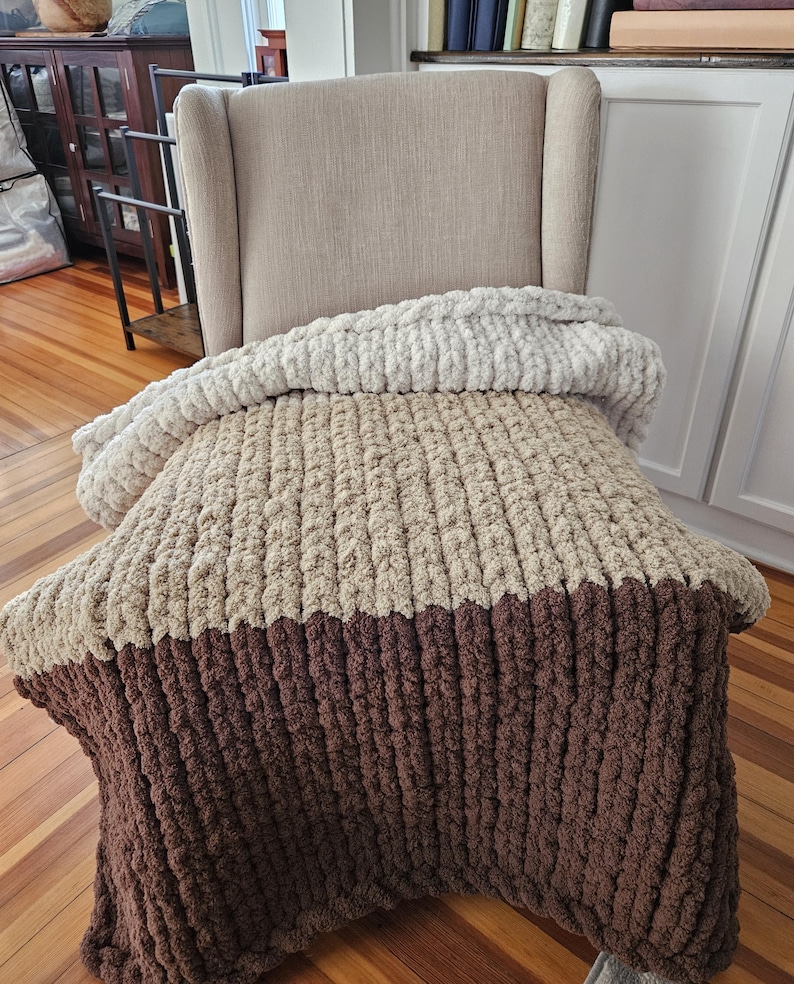 Pode incluir: Uma manta de malha grossa em tons de bege e castanho est&aacute; sobre uma poltrona bege. A manta apresenta um design texturizado e canelado, com uma sec&ccedil;&atilde;o superior bege claro, uma sec&ccedil;&atilde;o m&eacute;dia bege m&eacute;dio e uma sec&ccedil;&atilde;o inferior castanho escuro. A manta parece macia e aconchegante.