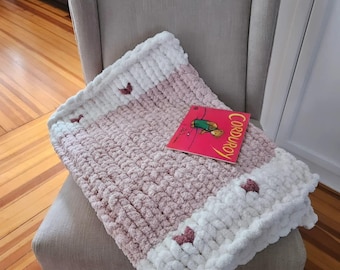 Pode incluir: Um cobertor de malha grossa rosa e branco com detalhes de coração e um livro intitulado "Corduroy". O cobertor está sobre uma poltrona bege, criando uma cena acolhedora e convidativa.