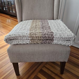 Pode incluir: Uma poltrona estofada em tons neutros com um cobertor de malha grossa sobre o assento. O cobertor apresenta listras brancas, bege e malva. A cadeira tem um encosto alto e pernas de madeira, criando uma est&eacute;tica aconchegante e convidativa.