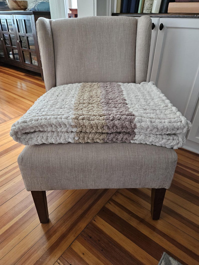 Pode incluir: Uma poltrona estofada em tons neutros com um cobertor de malha grossa sobre o assento. O cobertor apresenta listras brancas, bege e malva. A cadeira tem um encosto alto e pernas de madeira, criando uma est&eacute;tica aconchegante e convidativa.