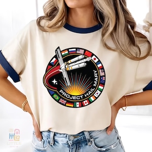 May include: Cream-coloured ringer tee with navy blue trim featuring a circular graphic. The graphic includes a rocket design, the text "PROJECT HAIL MARY," and flags of various nations. The shirt is a casual style.