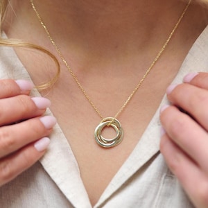 Peut inclure: Un collier doré avec un pendentif composé de trois anneaux entrelacés. Le collier est porté par une personne aux cheveux blonds et une chemise de couleur claire. Les anneaux sont d'une nuance d'or plus claire que la chaîne.