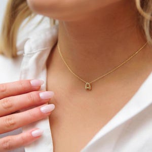 May include: A delicate gold-colored necklace featuring the letter "A" pendant. The pendant is a small, stylized capital letter. The necklace is worn around the neck, against a white shirt.