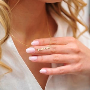 May include: A gold necklace with the word "nana" in a cursive font. The necklace is displayed against a white shirt. "Nana" is a term of endearment for grandmother.