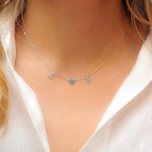 May include: A silver necklace featuring the letters "A" and "F" either side of a heart-shaped charm. The necklace is worn against a white shirt. The chain is fine and the letters are small, creating a subtle, elegant look.