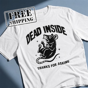 Peut inclure: Un t-shirt blanc à manches courtes avec un motif noir représentant un rat avec une croix sur un œil. Le texte au-dessus du rat indique DEAD INSIDE et le texte en dessous indique THANKS FOR ASKING. Le design inclut de petites étoiles.
