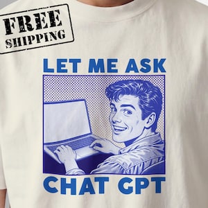 May include: A cream-colored t-shirt featuring a blue graphic print of a person using a laptop. The design includes a comic-style dot pattern and the text "LET ME ASK CHAT GPT" in bold blue capital letters, presented in a square frame.