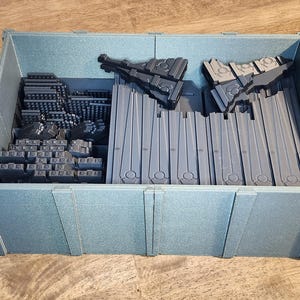 May include: A blue-gray plastic storage bin filled with various gray plastic components. The bin contains rectangular and geometric shapes, possibly for a model or construction project. The bin is on a wooden surface.