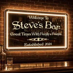 Puede incluir: Un letrero rectangular de LED montado en la pared con marco negro y luz blanca cálida. El texto dice: Welcome To Steve's Bar, Great Times With Family & Friends, Established 2024. El letrero presenta un diseño decorativo de filigrana.
