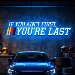 以下が含まれることがあります： 青いスポーツカーが、"IF YOU AIN'T FIRST, YOU'RE LAST"と書かれたネオンサインのあるガレージに駐車されています。ガレージには工具、オートバイ、レーシングヘルメットがあります。サインには黄色、赤、青のストライプがあります。