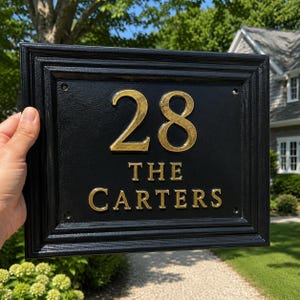 Peut inclure: Une plaque de maison rectangulaire noire avec un cadre en relief. La plaque présente le numéro 28 en doré en haut, suivi du texte THE CARTERS en lettres dorées. Il y a quatre petits trous de montage circulaires dans les coins.