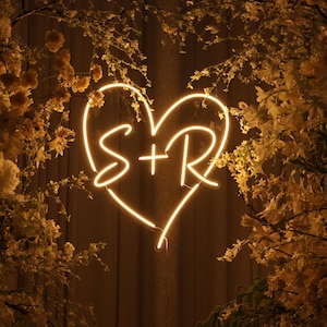 May include: A glowing neon heart sign with the letters "S + R" inside. The sign is surrounded by dried flowers and foliage, creating a warm, romantic ambiance. The neon light is a soft yellow color.