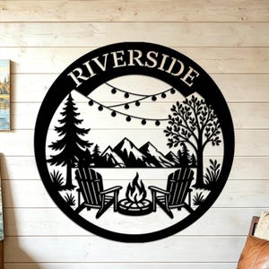 May include: A circular black metal wall sign featuring a cutout design of a campfire scene with two chairs, a pine tree, a deciduous tree, and mountains. The word RIVERSIDE is arched across the top, and a string of lights hangs above the scene.