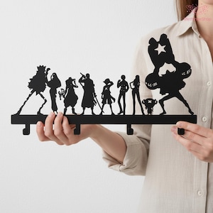 May include: A black metal wall hook featuring silhouettes of anime characters. The hook has several downward-facing hooks for hanging items. The characters are arranged in a row, with varying poses and details, creating a decorative and functional piece.