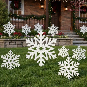 May include: White snowflake yard decorations of various sizes are displayed on a green lawn. The snowflakes are cut out with intricate designs and are placed in front of a house decorated for the holidays. The house has a brick exterior and is decorated with wreaths and lights.