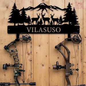 Peut inclure: Décoration murale en métal noir représentant des montagnes, des arbres et des silhouettes de cerfs, avec le nom "VILASUSO" en dessous. Deux arcs à poulies sont montés en dessous sur un mur en bois.